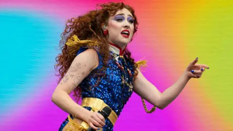 Getty Images Chappell Roan performs live on stage, holding a microphone in one hand and gesturing to the audience with the other. She wears bright purple eyeshadow, red lipstick and a blue sequined outfit with gold trim in front of a multi-colored backdrop.