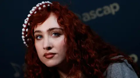 Getty Images Chappell Roan photographed at a red carpet event. Chappell is a woman in her twenties with long curly dyed red hair and blue eyes. She wears a beaded beret on top of her head and has dark red lipstick. 