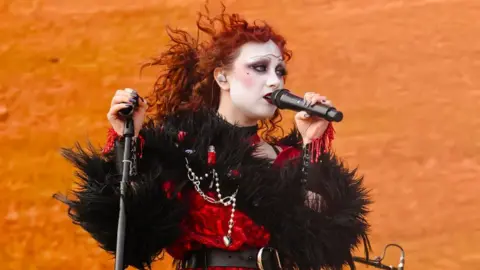 Getty Images Chappell Roan performs during the 2024 Boston Calling Music Festival at Harvard Athletic Complex on May 26, 2024 in Boston. Chappell has bright red curly hair and wears drag-style makeup, including a white face, heavy blush, and thin penciled eyebrows. She wears a red and black feathered dress and sings into a microphone in front of an orange backdrop. 